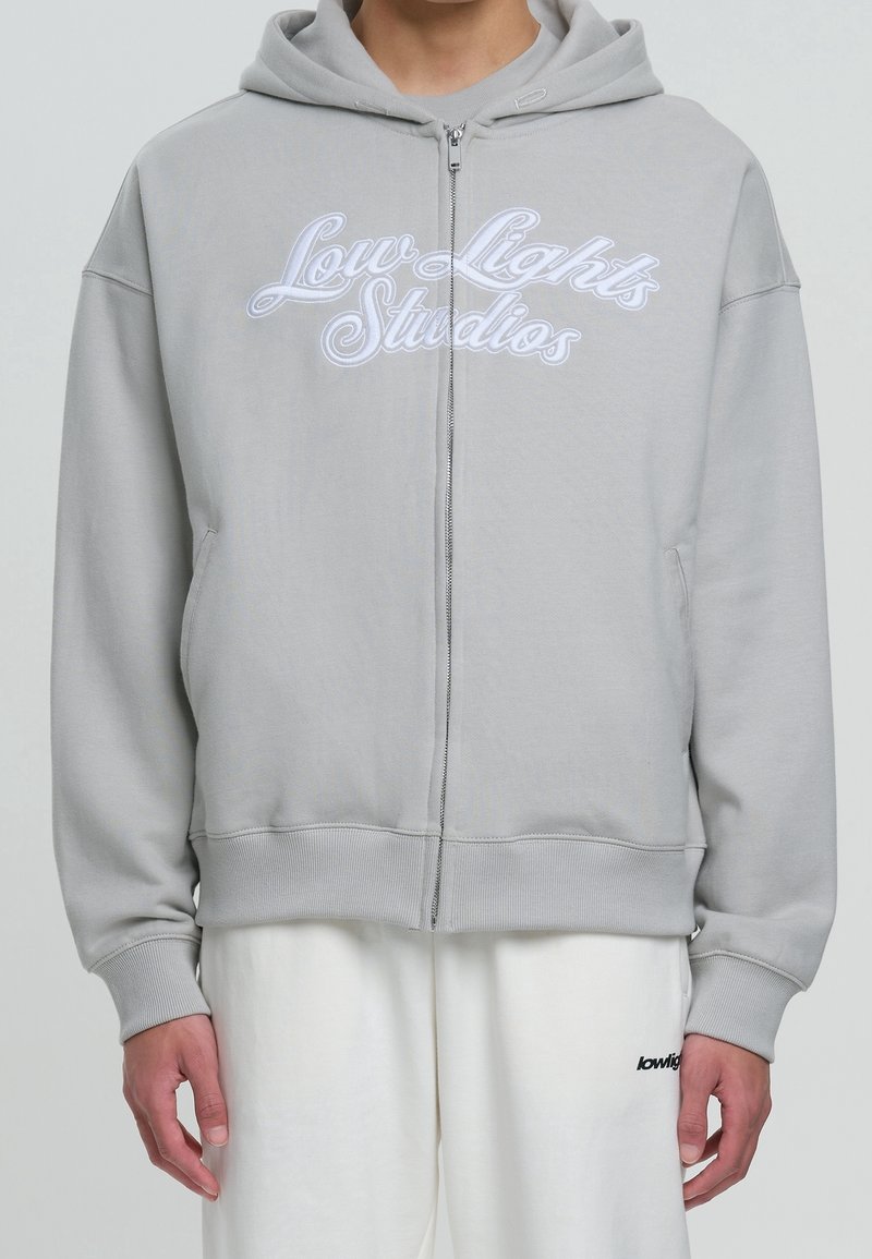 Grauer Kapuzenpullover mit Reißverschluss aus weichem Stoff, mit Vorderentaschen und weiß besticktem "Low Lights Studios" Schriftzug. Klassischer Schnitt mit Kapuze.