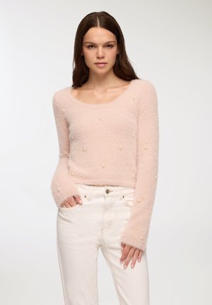 Maglione fluffy rosa chiaro con scollo rotondo, maniche lunghe e decorazioni simili a perle. Indossato con pantaloni in denim bianchi.