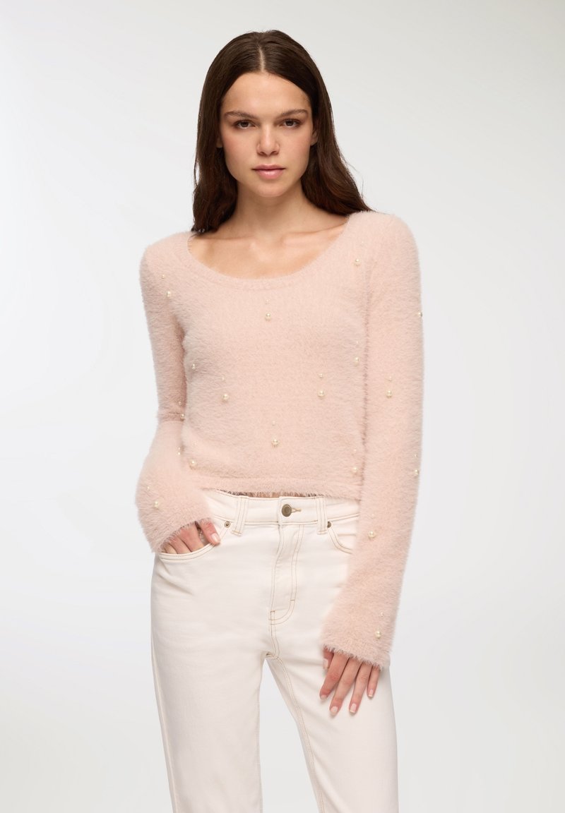 Jasnoróżowy puszysty sweter z okrągłym dekoltem, długimi rękawami i perłowymi zdobieniami. Noszony z białymi spodniami dżinsowymi.