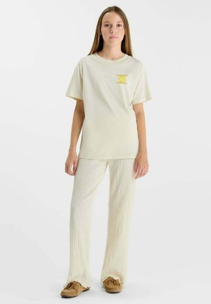 Jeune femme aux cheveux longs, vêtue d'un T-shirt beige, d'un pantalon texturé crème et de mocassins marron, debout sur un fond blanc.