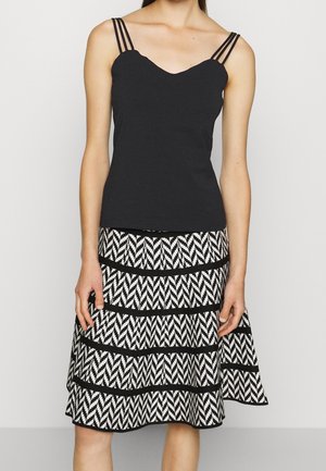 Femme portant un haut noir sans manches avec des bretelles triples et une jupe mi-longue noire et blanche à motifs en zigzag.