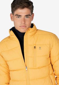 Chaqueta acolchada amarilla con textura acolchada, cuello alto, cierre delantero con cremallera y un pequeño bolsillo en el pecho con un logo en el lado izquierdo.