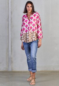 Bunte, gemusterte Bluse mit pinken Fächer- und geometrischen Drucken, leichtem Stoff, langen Ärmeln und V-Ausschnitt; kombiniert mit hochgekrempelten Denim-Jeans.