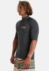 Grå rash guard med kort ärm och hög krage samt en färgglad grafisk logotyp. Bärs med ljusa shorts med horisontella ränder i grön, orange och vit.