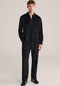 Camicia nera a bottoni con un tessuto texturizzato e colletto aperto, abbinata a pantaloni neri e sneakers neri. Design semplice e moderno.