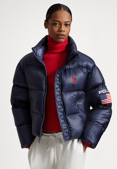 Puffer jacket in navy blue with a shiny texture, featuring a red logo and patch on the sleeve, paired with a red turtleneck underneath.
