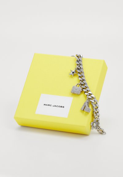 Silverfärgad charm-armband med stjärna, hänglås, högklackade skor och blommor som detaljer på en gul presentlåda med etikett "Marc Jacobs".