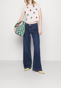 Top de ganchillo con bordado de insectos multicolores, jeans de mezclilla de tiro alto y acampanados, bolsa a cuadros verde y rosa, y zapatos amarillos de punta.
