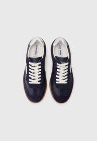 Marineblå sneakers lavet af ruskind og læder, med hvide snørebånd, en tekstureret sål og et brandmærke på tungen.