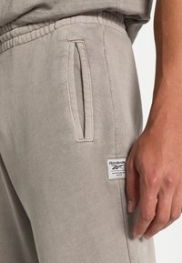 Beige Reebok-träningsbyxor med elastisk midja, sidoficka med slits, etikett i naturlig färg och en persons högra hand som vilar vid höften.