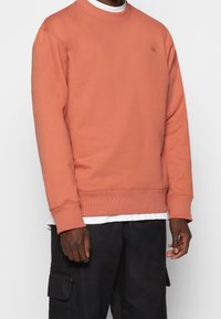 Uomo che indossa un maglione rosa salmone sopra una camicia bianca e pantaloni cargo neri su uno sfondo semplice.