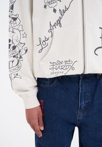 Sudadera con cremallera beige con diseños gráficos en negro y texto, combinada con jeans azul oscuro que tienen un acento rojo en el bolsillo.