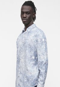 Šviesiai mėlyna, ilgomis rankovėmis su raštais marškiniai su užsegamu apykakliu, puošti gėlių motyvais ir krūtinės kišene. Medvilnės audinys, laisvas modelis.