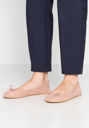 Personne portant un pantalon bleu marine et des ballerines en suède rose clair avec de petits nœuds, se tenant sur un sol blanc lisse.
