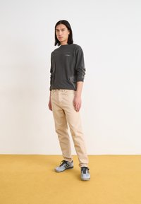 Gray long-sleeve shirt with a subtle logo, beige jeans, and gray sneakers. The setting features a yellow floor and neutral background.