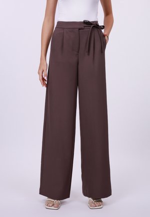 Frau trägt hochtaillierte, weit geschnittene braune Hosen mit gebundenem Gürtel, weißes Tanktop und weiße offene Sandalen vor einfachem Hintergrund.