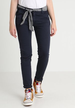 Persoon draagt donkere marineblauwe broek met een vastgeknoopte riem van patroonstof en kleurrijke hoge sneakers met witte veters.