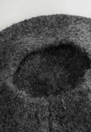 Gekrulde, gestructureerde donkergrijze wollen baret met fuzzy, lusvormige vezels tegen een lichte achtergrond.