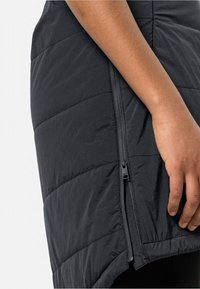 Zwarte geïsoleerde vest met een puffy textuur, voorzien van een zijrits en een diagonale zoom. Close-up van een hand die op de stof rust.