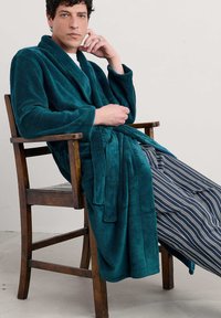 Tirkizna plišana kupaća haljina s širokim rukavima, vezana u struku, s prednjim džepovima, u kombinaciji s plavim i bijelim prugastim hlačama. Sjedi na drvenoj stolici.