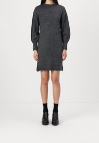 Robe en tricot gris avec un col montant, des manches bouffantes et des fentes latérales. Le tissu semble doux et chaud. Portée avec des chaussures et des chaussettes noires.