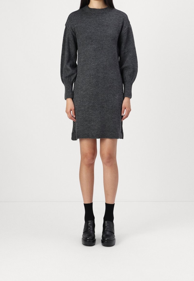 Robe en tricot gris avec un col montant, des manches bouffantes et des fentes latérales. Le tissu semble doux et chaud. Portée avec des chaussures et des chaussettes noires.