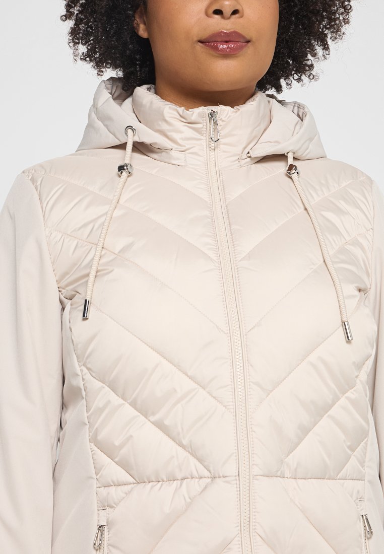 Hellbeige gepolsterte Jacke mit Kapuze, ausgestattet mit einer Zickzack-Steppung, Reißverschluss und Kordelzügen. Glatte Textur mit kontrastierendem Ärmelmaterial.