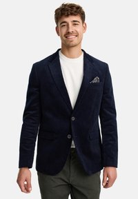 Blazer en velours côtelé bleu marine avec une coupe ajustée, deux boutons, deux poches avant et un mouchoir de poche à motifs. Porté sur une chemise blanche.