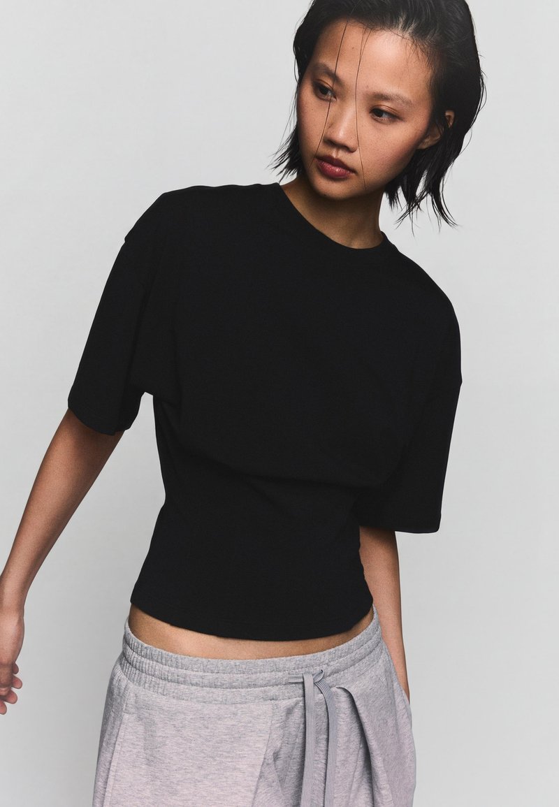Femme portant un haut ample noir à manches courtes rentré dans un pantalon de jogging gris clair avec un cordon, regardant vers le bas et légèrement sur le côté.