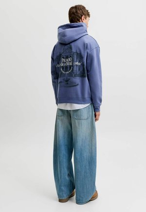 Personne portant un sweat à capuche bleu avec une illustration de palais et un texte dans le dos, associé à un jean bleu clair à jambes larges et des chaussures marron.