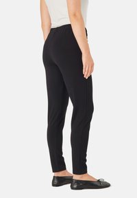 Zwarte stretchbroek met een elastische tailleband, slim-fit enkel lengte ontwerp, gecombineerd met platte zwarte schoenen, gezien vanaf de zijkant.