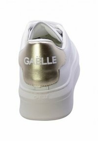 Sneaker bianca con un accento in oro metallico sul tallone, con logo "GAËLLE", superficie liscia, punta rotonda e suola ammortizzata.