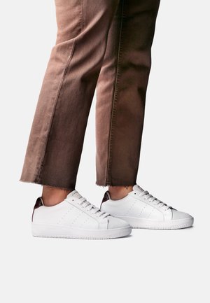 Personne portant un pantalon marron raccourci et des baskets blanches basses avec un détail talon bordeaux, debout sur un fond clair.