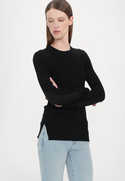 Black ribbed knit sweater with long sleeves, featuring side slits and black panel accents on shoulders. Styled with light blue jeans.