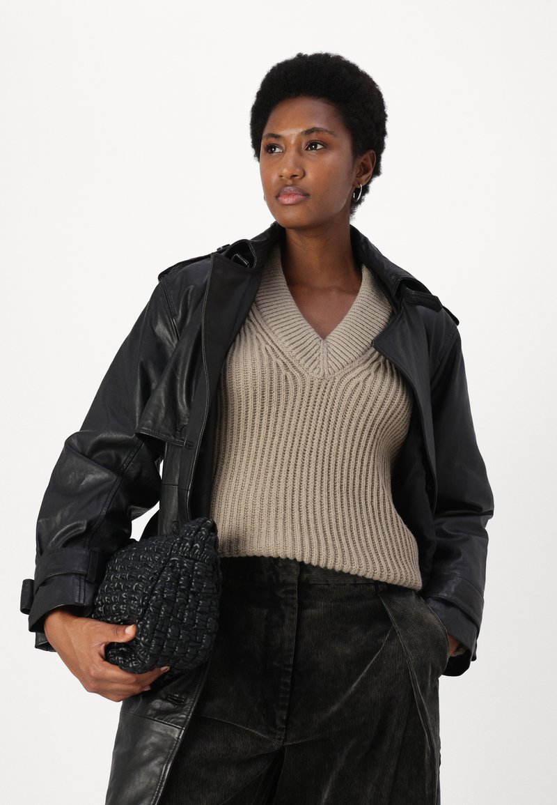 Schwarzer Leder-Trenchcoat über einem beigen, gestrickten V-Ausschnitt-Pullover, kombiniert mit dunklen Cordhosen und einer strukturierten schwarzen Handtasche.