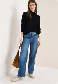 Czarny sweter z fakturą dzianiny, szerokie niebieskie dżinsy, beżowe sneakersy z niebieskimi akcentami oraz brązowa zamszowa torba na ramię.