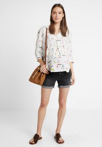 Blusa branca com padrões coloridos, mangas três-quartos e um detalhe em botão, combinada com calções de ganga escuros e uma mala de ombro castanha.