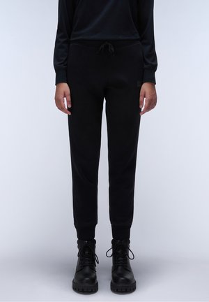 Person wearing black jogger pants with drawstring and black lace-up ankle boots, standing against a plain light background.