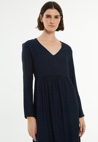 Vestido a cuadros en azul marino con mangas largas, escote en v y cintura fruncida, fabricado con una tela suave para un ajuste cómodo.