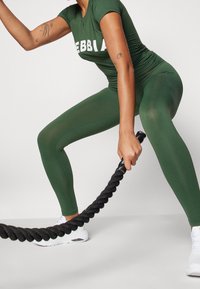 Zelený sportovní outfit s těsným tričkem, na kterém je výrazný bílý text. Legíny mají hladkou, pružnou texturu. Bílá sneakers a černá cvičební lano dotvářejí celkový vzhled.