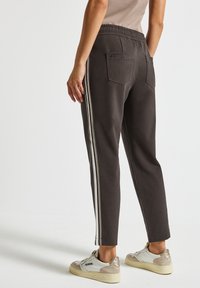 Person, der dunkelgraue Jogginghosen mit zwei hellen Seitenstreifen, Gesäßtaschen und beige-weißen Freizeitsneakern vor einem weißen Hintergrund trägt.