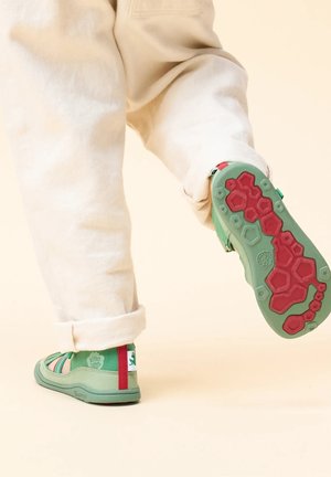 Green casual shoes with varied straps, textured rubber sole in green and red, with a logo accent on the heel. Worn with beige pants.