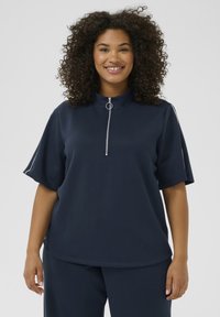 Navy blue short-sleeve top with a high neck, zipper, and circular hardware detail. Fabric appears smooth with a relaxed fit and side accents.