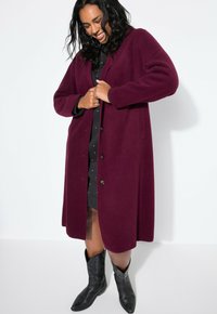 Cappotto in lana bordeaux con una vestibilità rilassata, maniche lunghe e chiusura a bottoni. Indossato sopra un vestito nero e abbinato a stivali da cowboy neri.