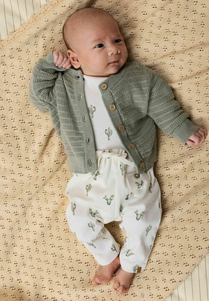 Niemowlę leżące na beżowym teksturowanym kocu, ubrane w zielony dziany sweterek i białe spodnie z zielonymi nadrukami kaktusów.