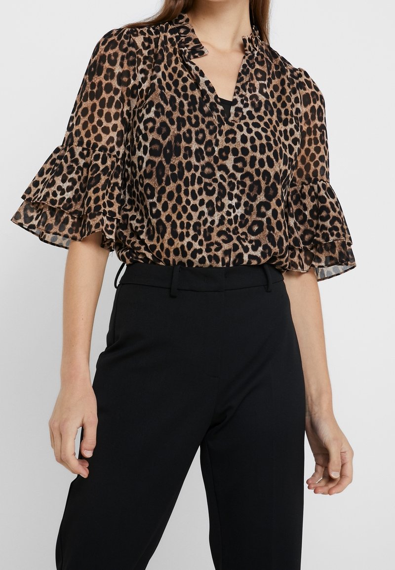 Femme portant un chemisier à imprimé léopard avec des manches courtes à volants, associé à un pantalon noir taille haute, debout devant un fond uni.