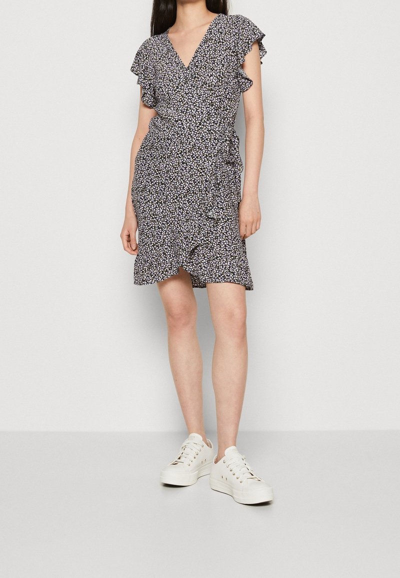Femme portant une robe portefeuille courte noire à motifs floraux avec des manches et un ourlet à volants, associée à des baskets blanches, debout devant un fond uni.