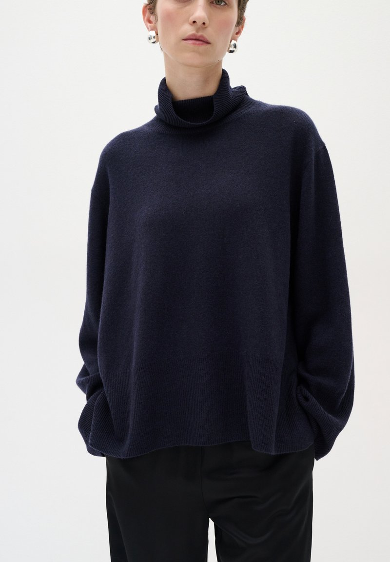 Tumši zili, pārmērīgi lieli turtlenecks džemperis ar ribotām aprocēm un apakšmalu, izgatavots no mīksta materiāla, kombinēts ar melnām biksēm.