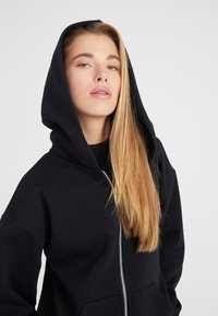 Sudadera con cremallera negra y capucha grande, fabricada en un tejido suave. Presenta una cremallera plateada y bolsillos delanteros, diseñada con un ajuste relajado.