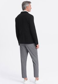 Schwarzer Blazer mit taillierter Passform, graue Hose und weiße Sneaker. Der Blazer hat einen Reverskragen und vier Knöpfe an jedem Ärmel.